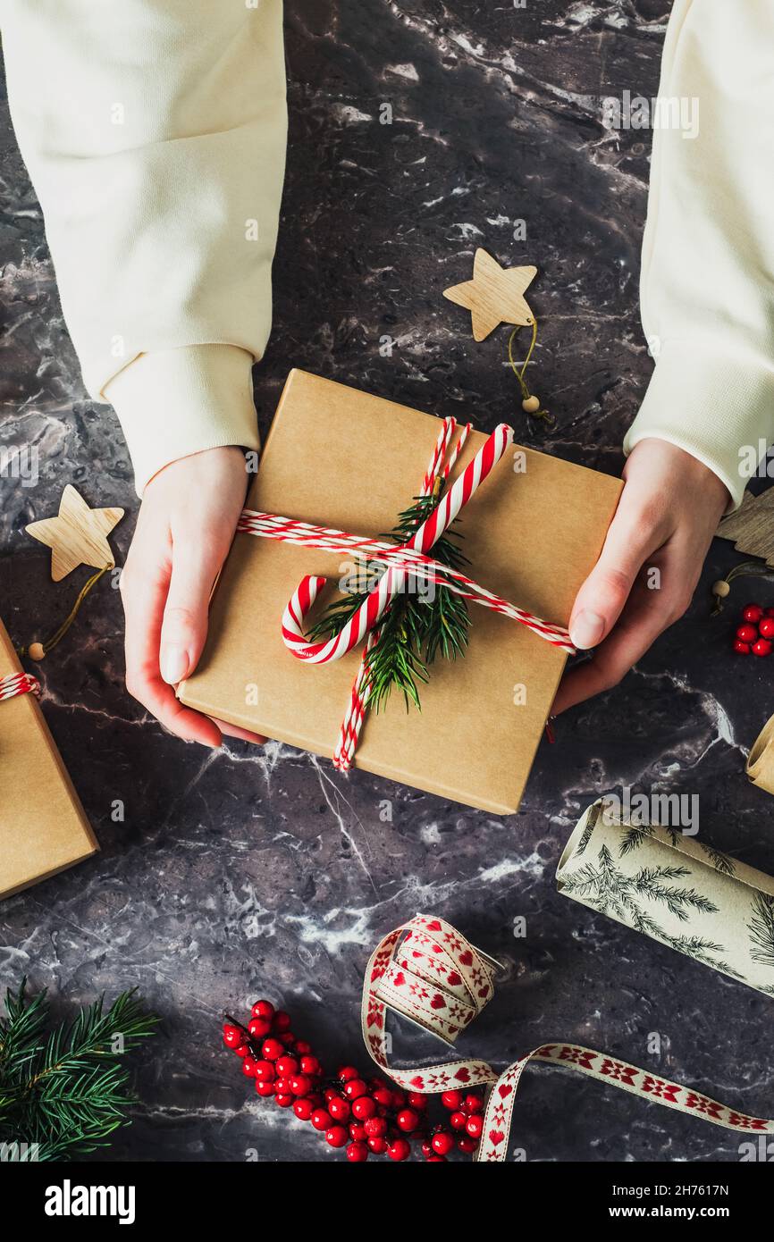 Womans hand holding Christmas gift box over slate desk table. Vintage Christmas present decorated red and white striped ribbon, fir branch, candy cane Stock Photo
