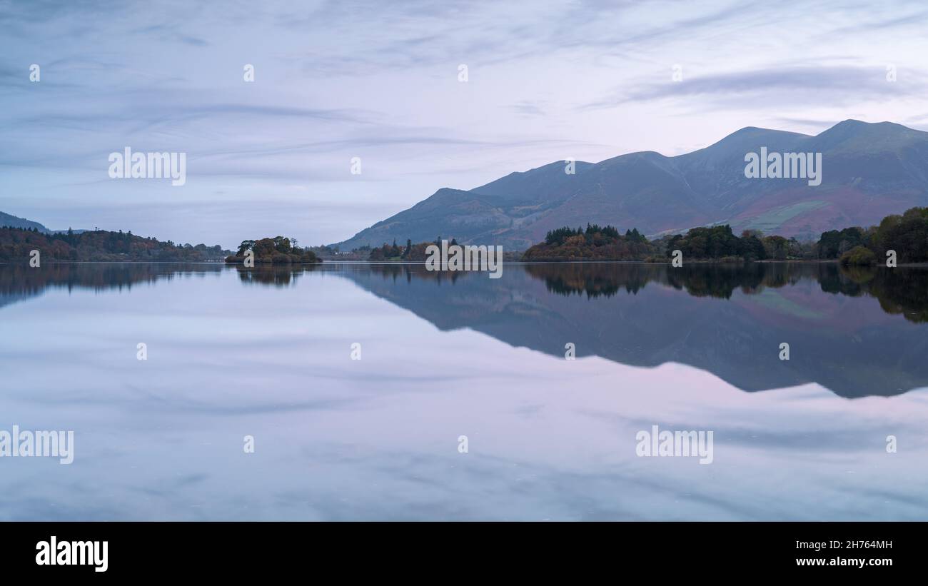 The Skiddaw Massif is reflected in a perfectly still Derwentwater during a subtle pastel sunrise in autumn, with wispy clouds framing the scene. Stock Photo