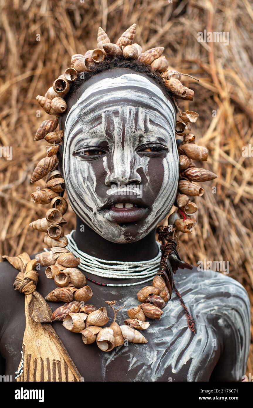 young girl from the Mursi tribe with white face painting and seashells around her head and neck in front of a grass hut Stock Photo