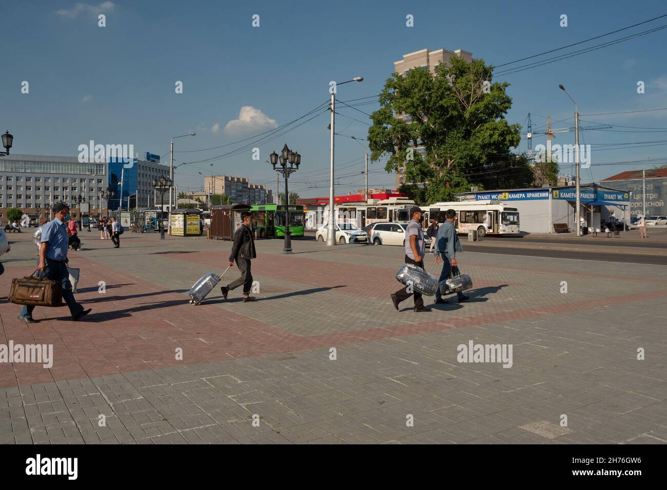 Migrants from the southern republics walk along the station square with their luggage to a bus stop on a sunny summer day. Stock Photo