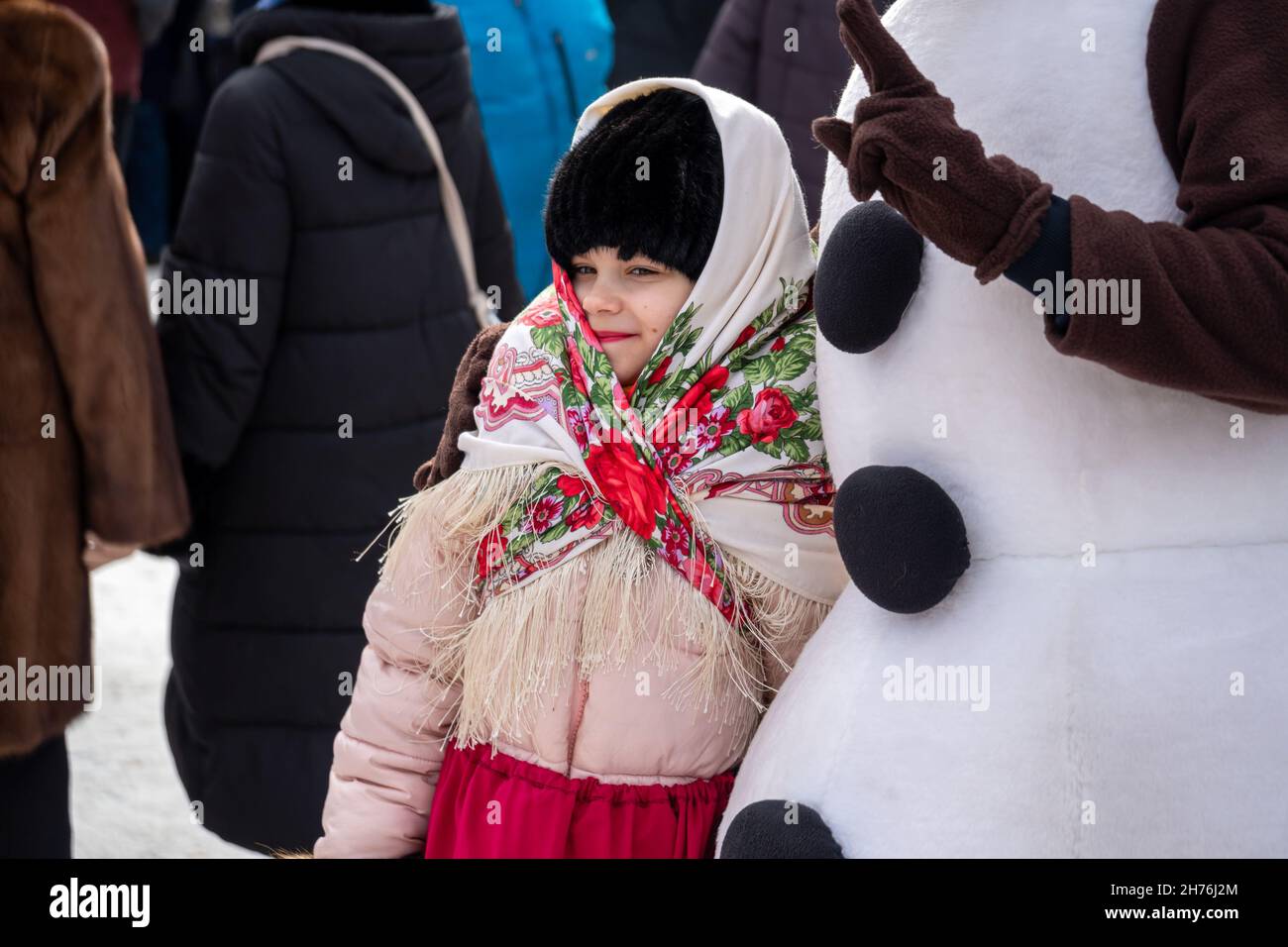 A girl in a Russian folk headscarf stands near a snowman actor at the Farewell to Winter festival in a city park. Stock Photo