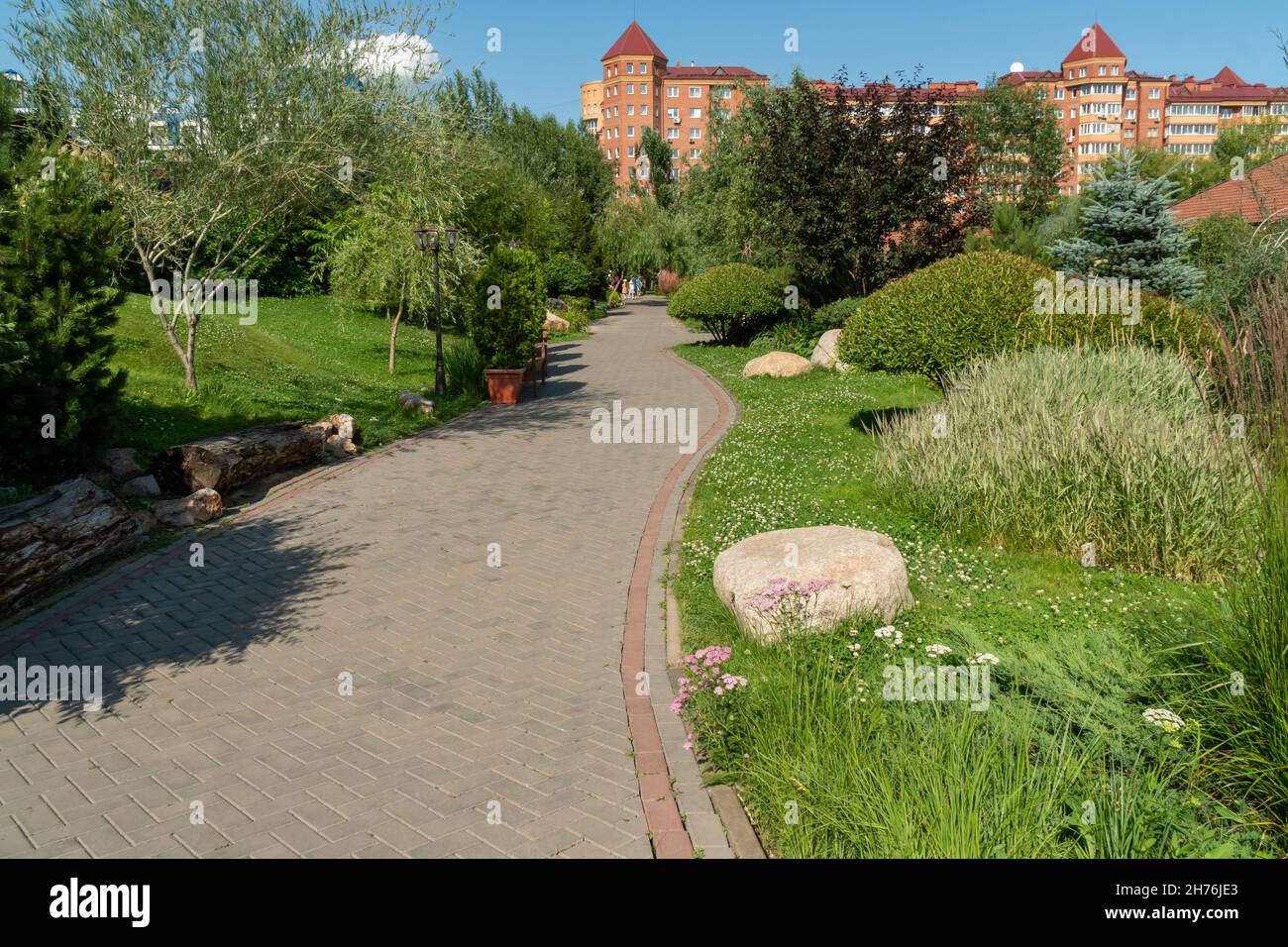 A flexible path between ornamental plants leads through the park to residential buildings on a sunny summer day. Stock Photo