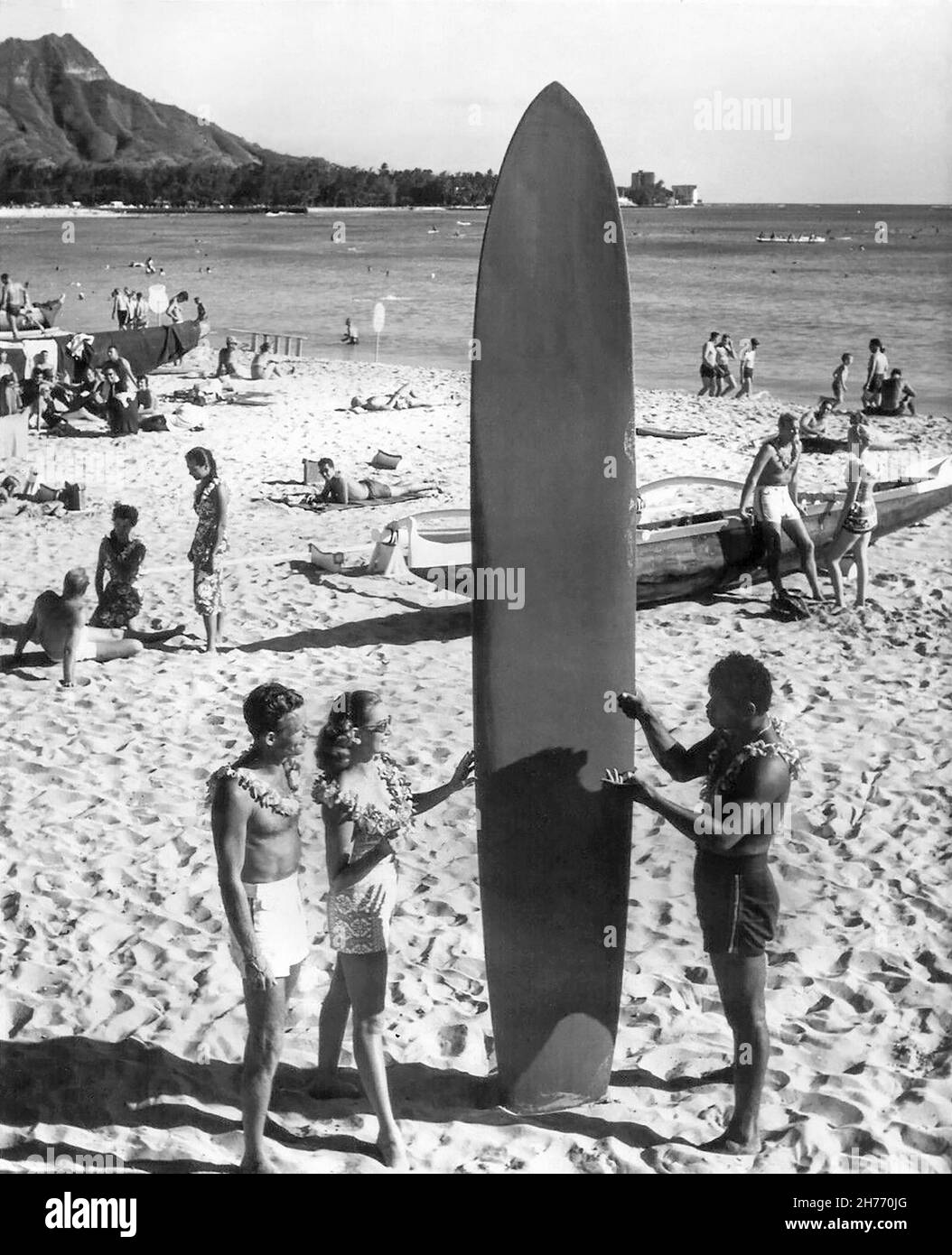 Vintage Waikiki Beach, Hawaii, travel photo from the 1960s of tourists with a Hawaiian surfer holding a wooden longboard, with a beached outrigger canoe and Diamond Head in the background. (USA) Stock Photo