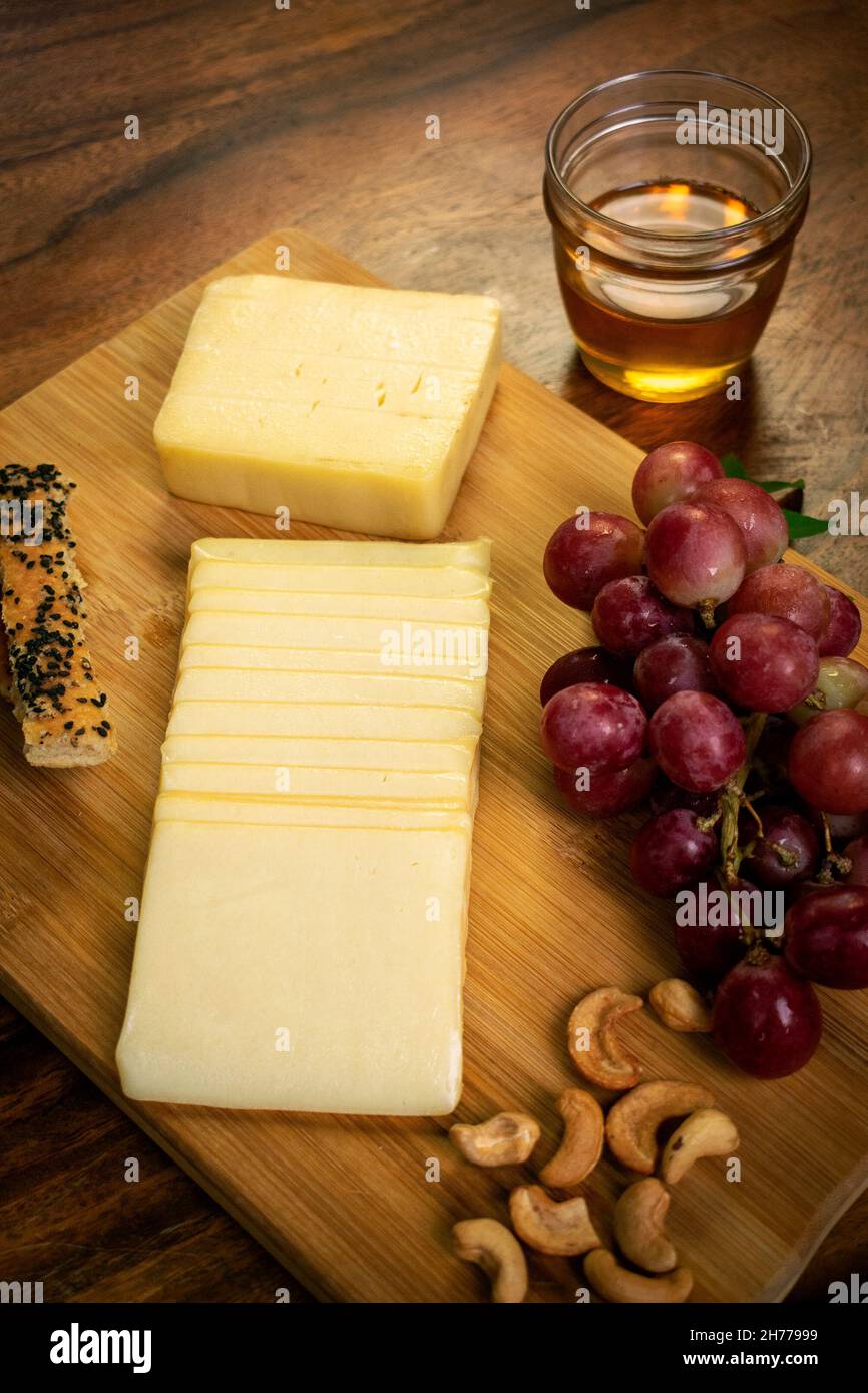 Sliced cheddar cheese block on wood board with grapes, cashew nuts, bread sticks and honey Stock Photo