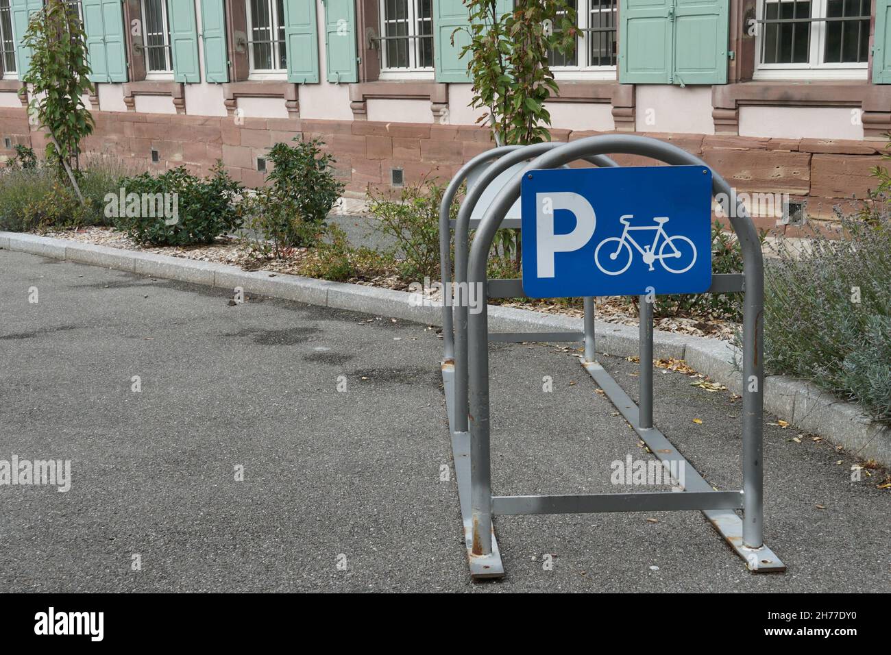 Arch metal frame for parking bicycles with blue sign containing letter P and pictogram of bicykle situated on the street near green house. Stock Photo