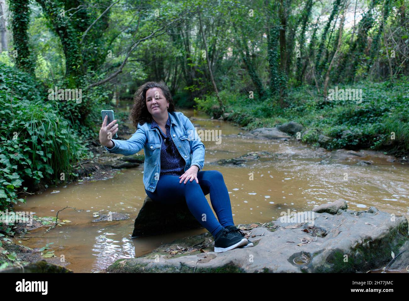 Woman sitting taking a selfie next to a stream running through a gallery forest. Stock Photo