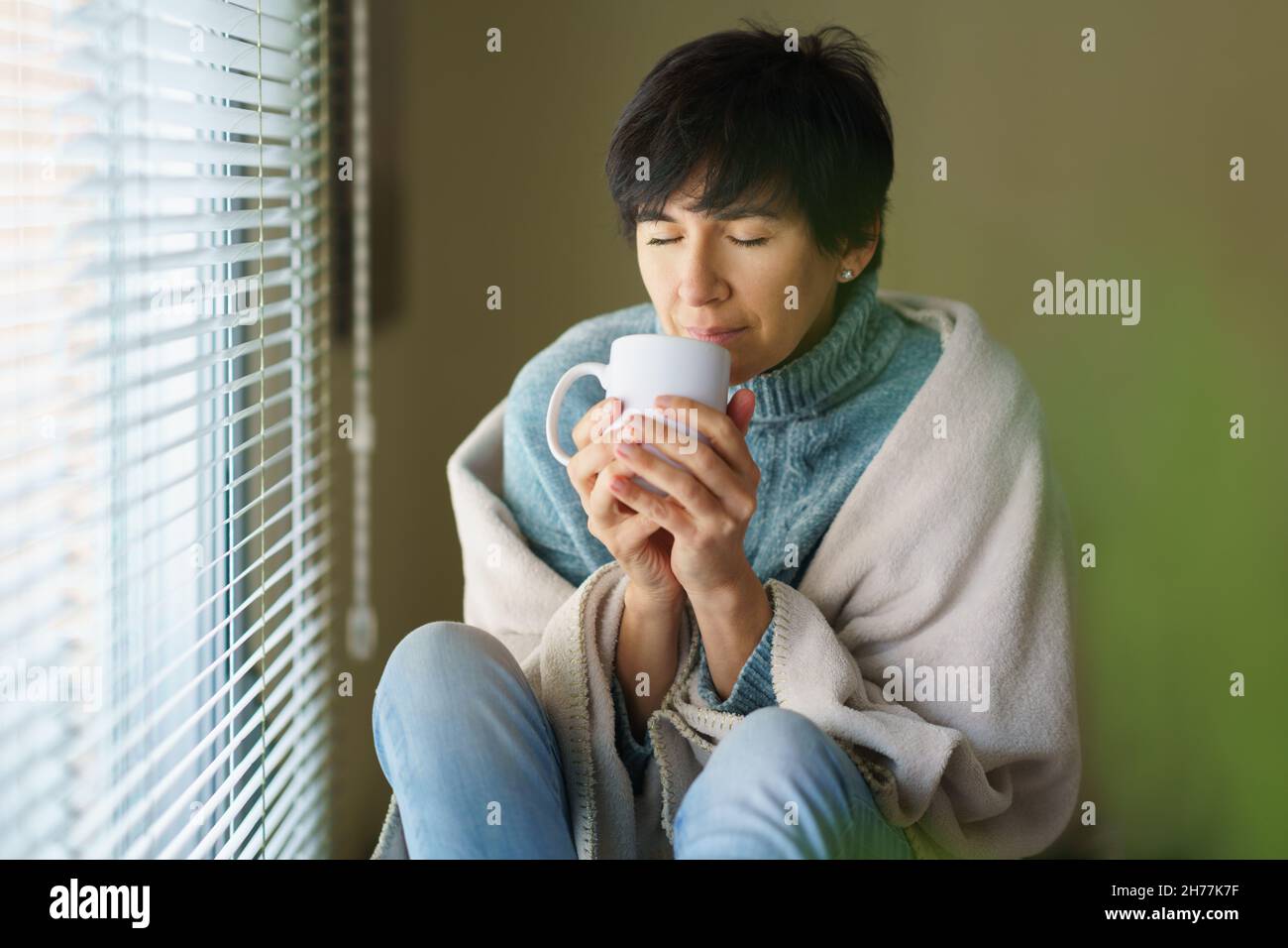 Middle-aged woman smelling coffee from her cup near a window. Stock Photo