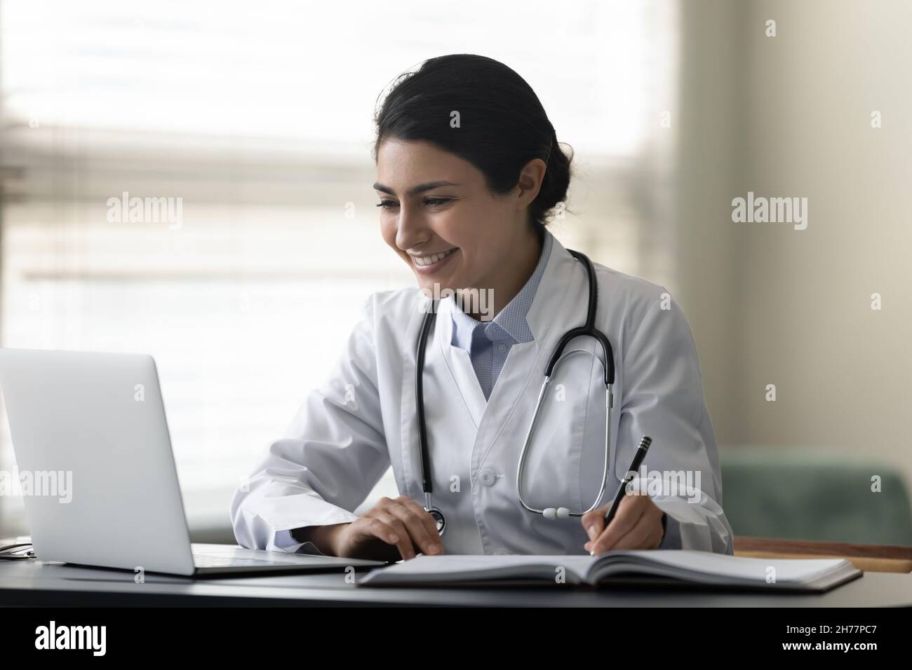 Smiling Indian female doctor physician using laptop, taking notes Stock Photo
