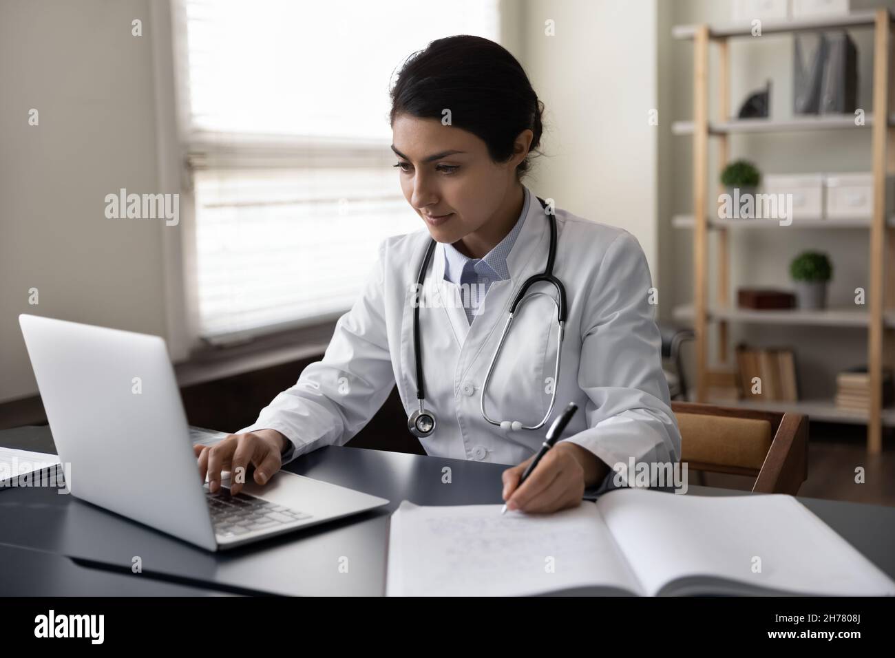 Serious Indian woman doctor physician using laptop, taking notes Stock Photo