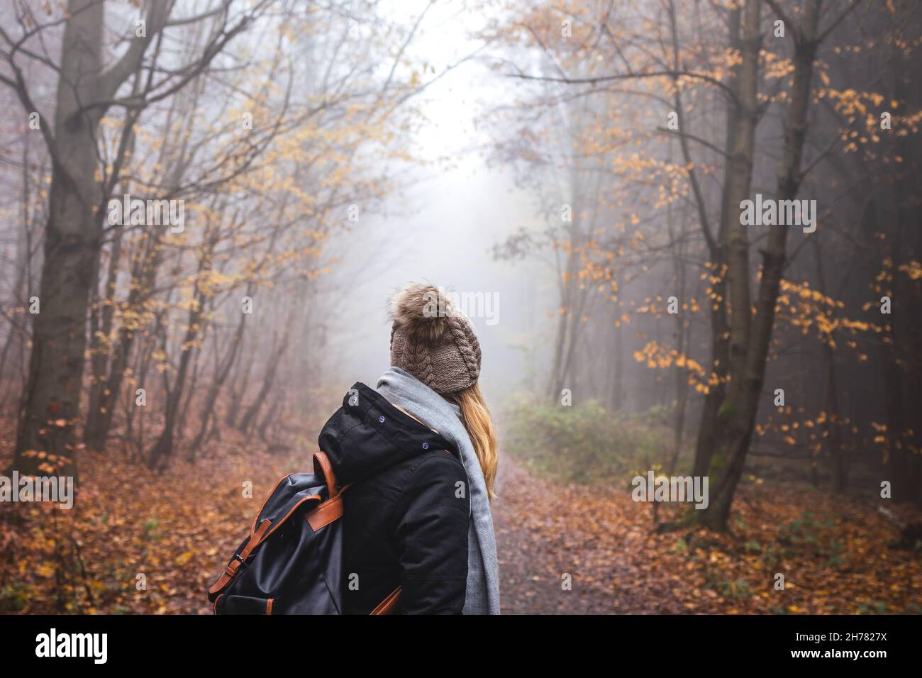 Woman with backpack and knit hat hiking in foggy forest. Hiker looking for adventure in nature. Mystery woodland Stock Photo