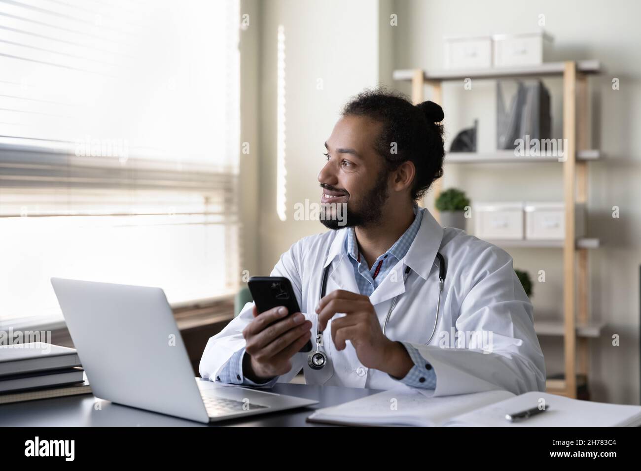 Smiling pensive African American doctor holding smartphone, sitting at desk Stock Photo