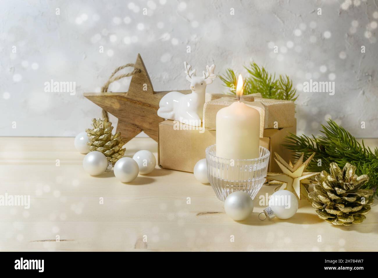 Christmas decoration with a warm white candle, porcelain deer, baubles, wooden stars and fir branches against a light gray background with snowy bokeh Stock Photo