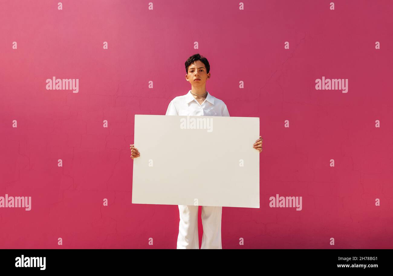 Young queer activist holding a white banner against a pink background. Assertive teenage boy displaying a blank placard in a studio. Young gay boy com Stock Photo