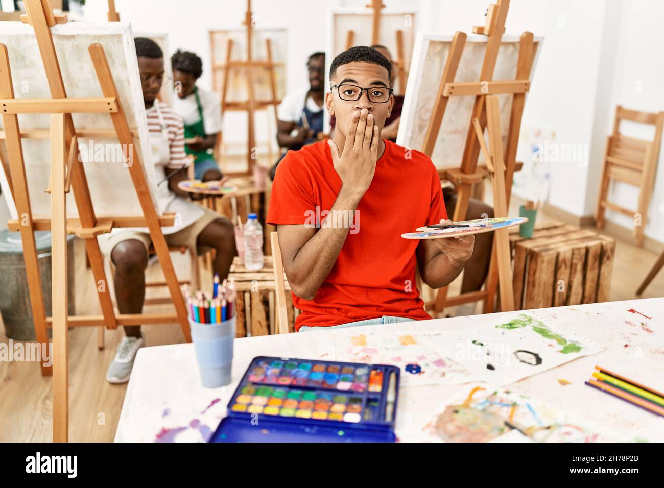 Young african man at art studio covering mouth with hand, shocked and ...