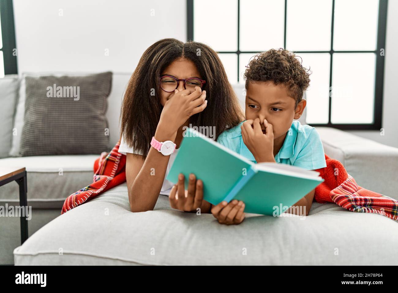 Two siblings lying on the sofa reading a book smelling something stinky and disgusting, intolerable smell, holding breath with fingers on nose. bad sm Stock Photo