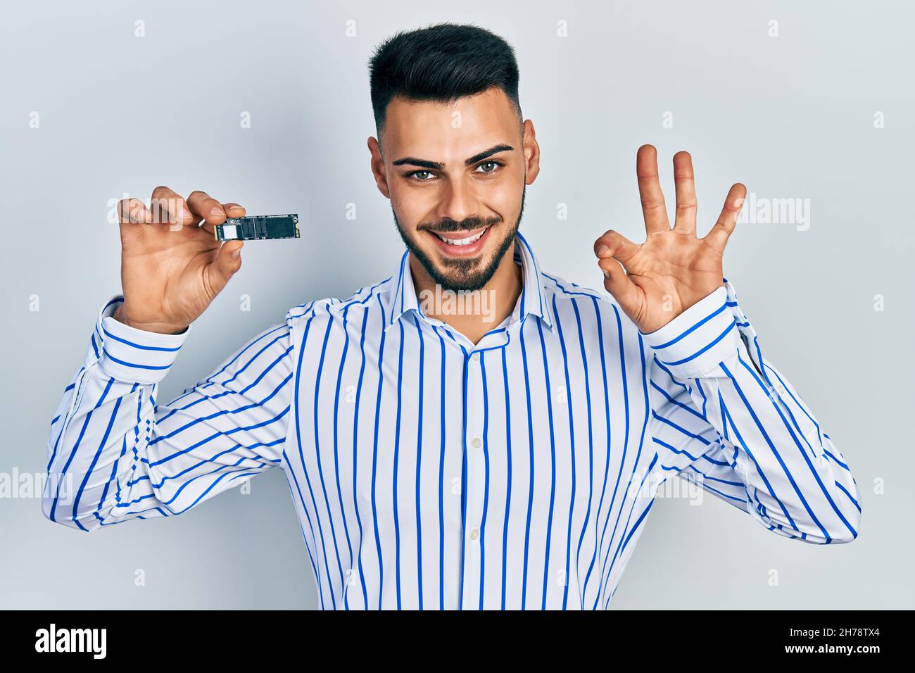 Young hispanic man with beard holding ssd memory doing ok sign with ...
