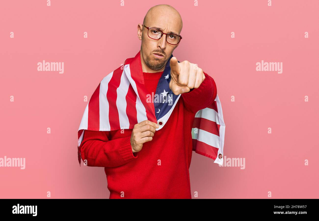 Bald man with beard wrapped around united states flag pointing with finger to the camera and to you, confident gesture looking serious Stock Photo