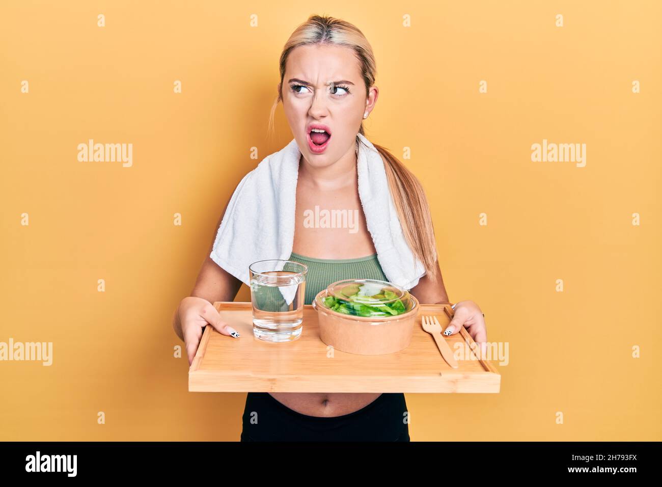 Young blonde girl wearing sportswear eating healthy food angry and mad