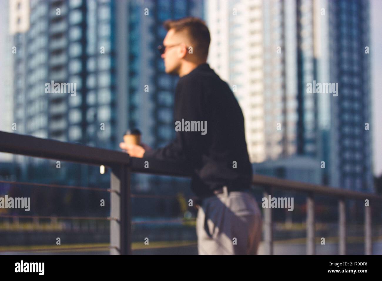 Unrecognizable young man in business suit is leaning against a metal railing on a pier against multistory apartment buildings. Pensive, reflective, in Stock Photo