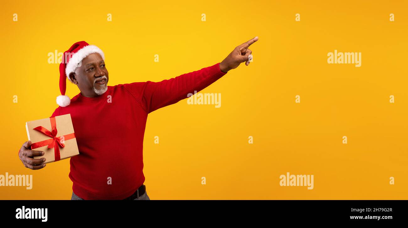 Merry Christmas. Senior black man holding Xmas gift, pointing aside at empty space for your ad on orange background Stock Photo
