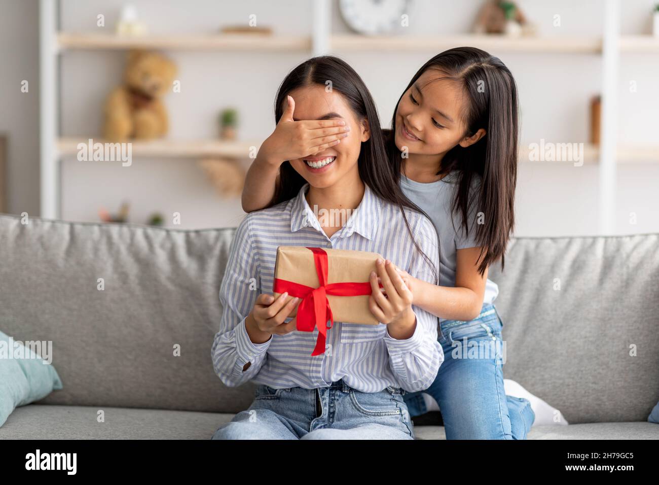 https://c8.alamy.com/comp/2H79GC5/playful-asian-girl-making-surprise-for-her-happy-mother-closing-her-eyes-and-giving-gift-box-while-sitting-on-couch-2H79GC5.jpg