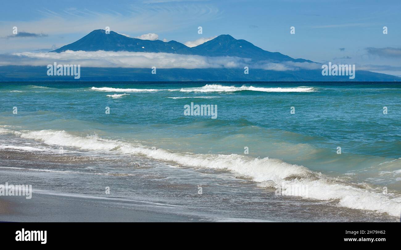 The emerald waves of the Sea of Okhotsk against the backdrop of active volcanoes Bogdan Khmelnitsky and Chirp on the island of Iturup are truly great. Stock Photo