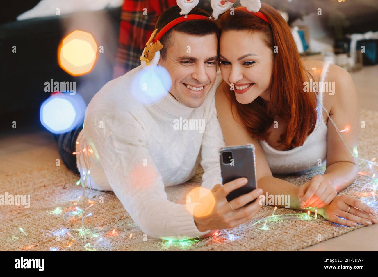 a happy married couple is lying on the floor at home near the Christmas tree and taking pictures of themselves. Stock Photo