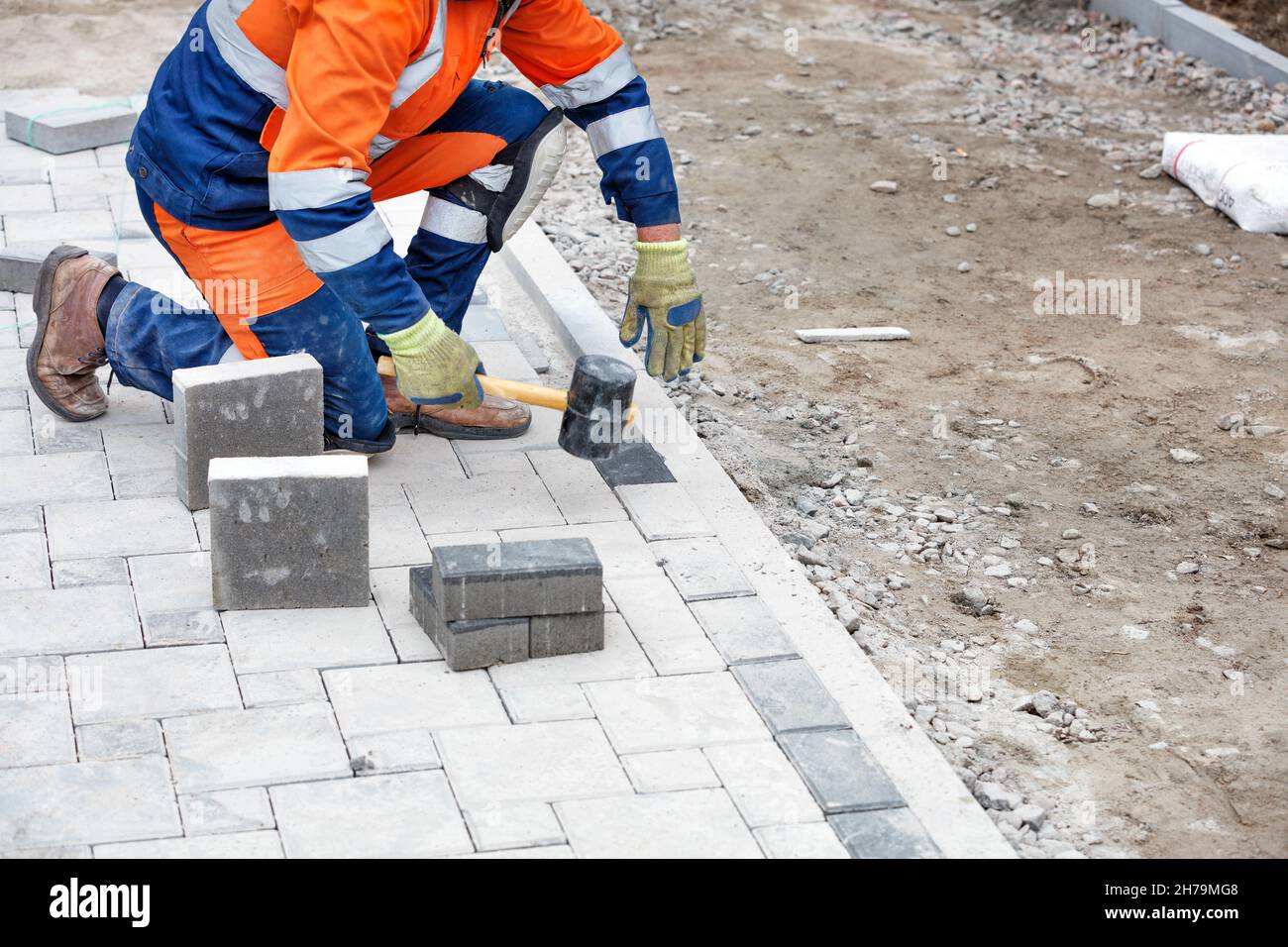 A bricklayer in a blue and orange overalls squatting down paving slabs