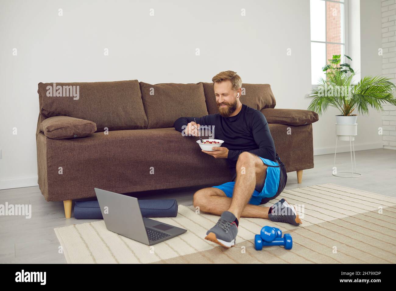 Man having break during fitness workout at home, eating healthy food and using laptop Stock Photo