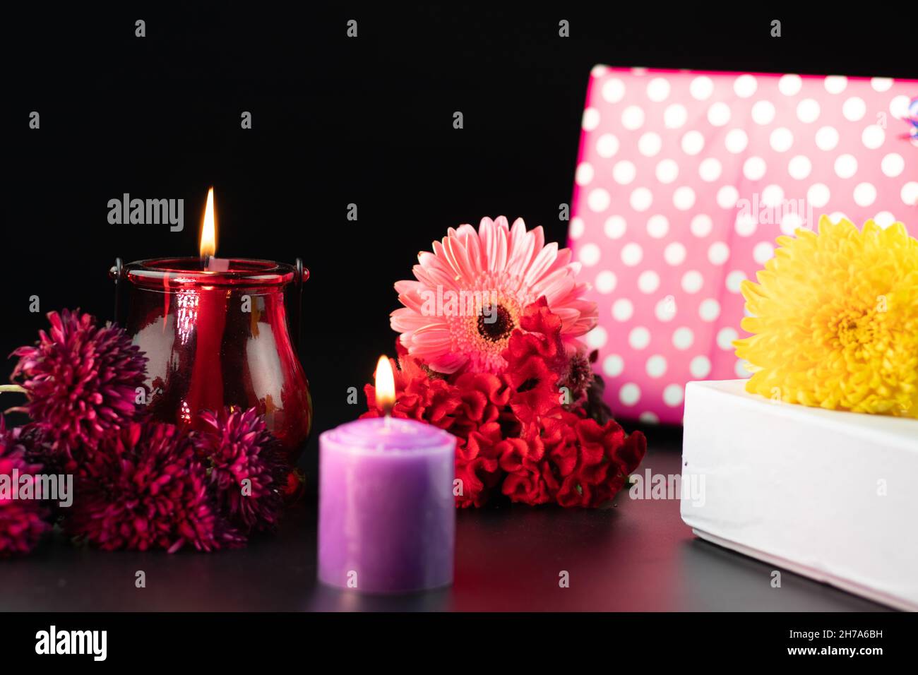 Selective Focus On Decorative Red Or Maroon Pillar Wax Candle Glowing In Designer Glass Holder Lantern. Beautiful Fresh Colorful Flowers And Gift Boxe Stock Photo