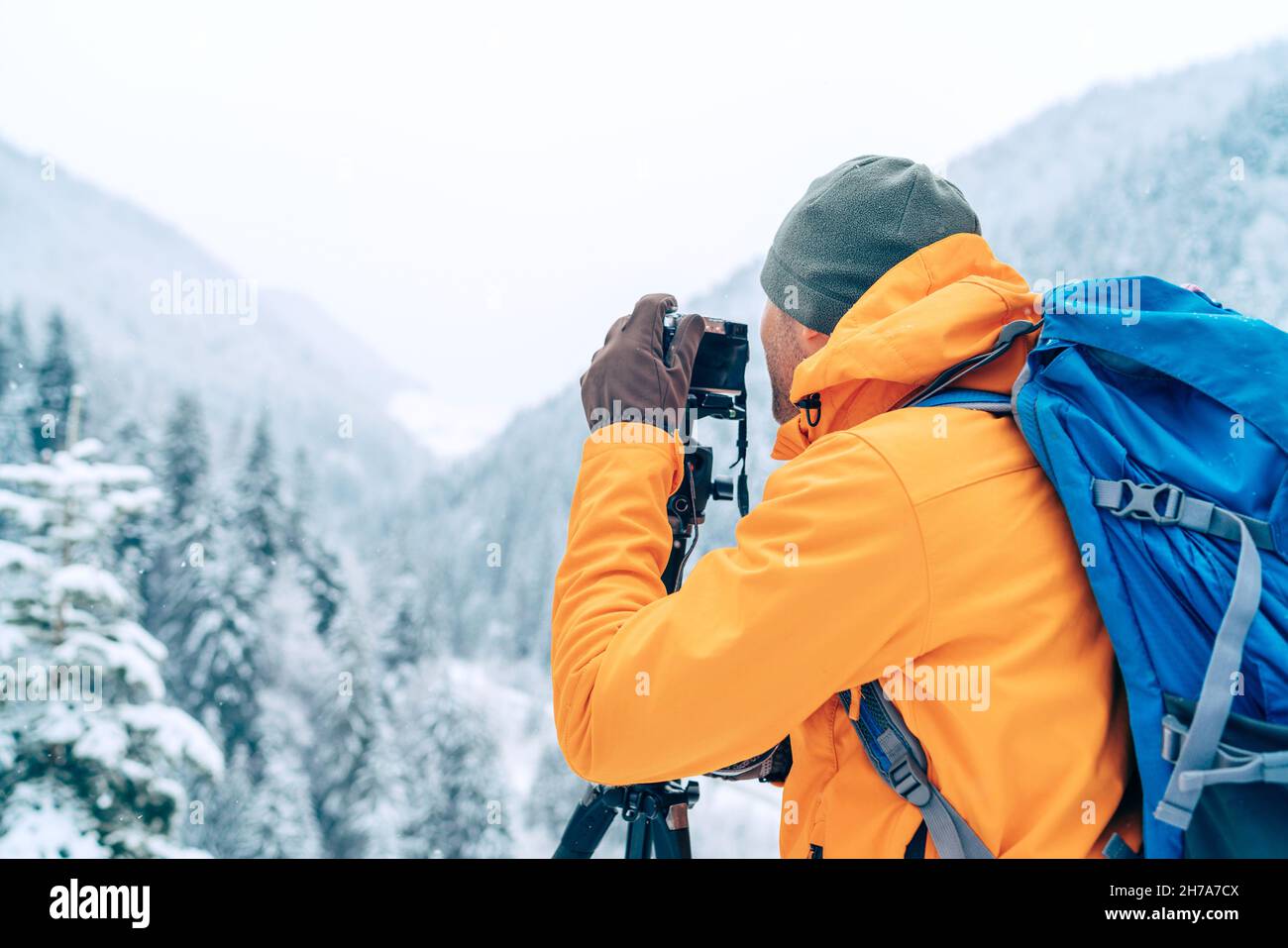 Photographer dressed orange softshell jacket with backpack making a landscape shoot using a digital camera and tripod while he trekking winter mountai Stock Photo