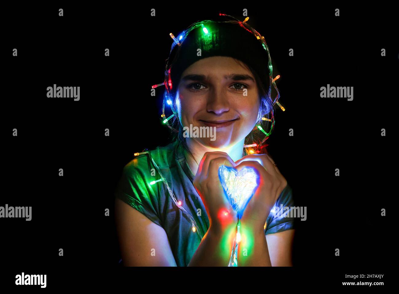 Portrait of a festive young woman with garland on her head and body. Bokeh and lights. Girl 25 years old in a hat smiles in the dark and show heart by Stock Photo
