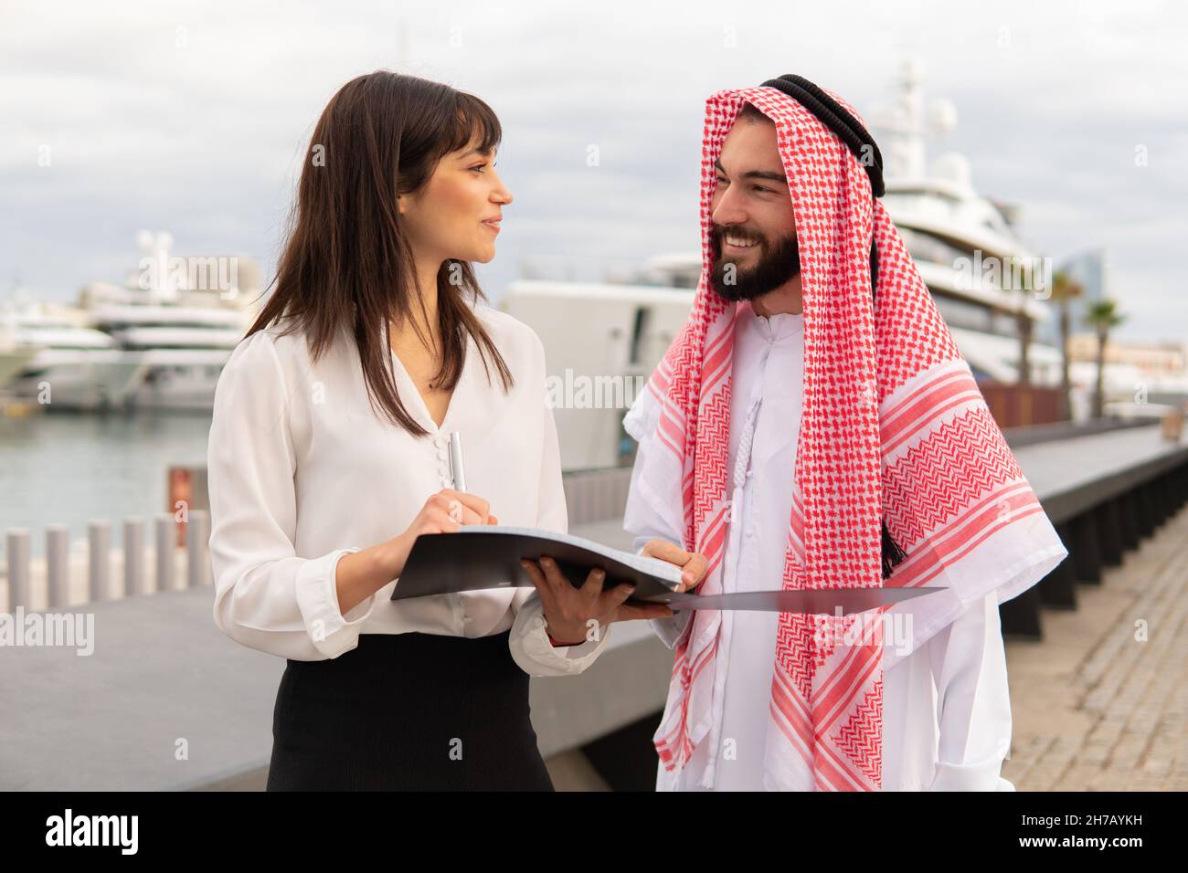 Young friendly europian businesswoman explaning contract details to arab man client during outdoor meeting in port, waiting saudi male business partner to sign document. Diverse partnership concept Stock Photo