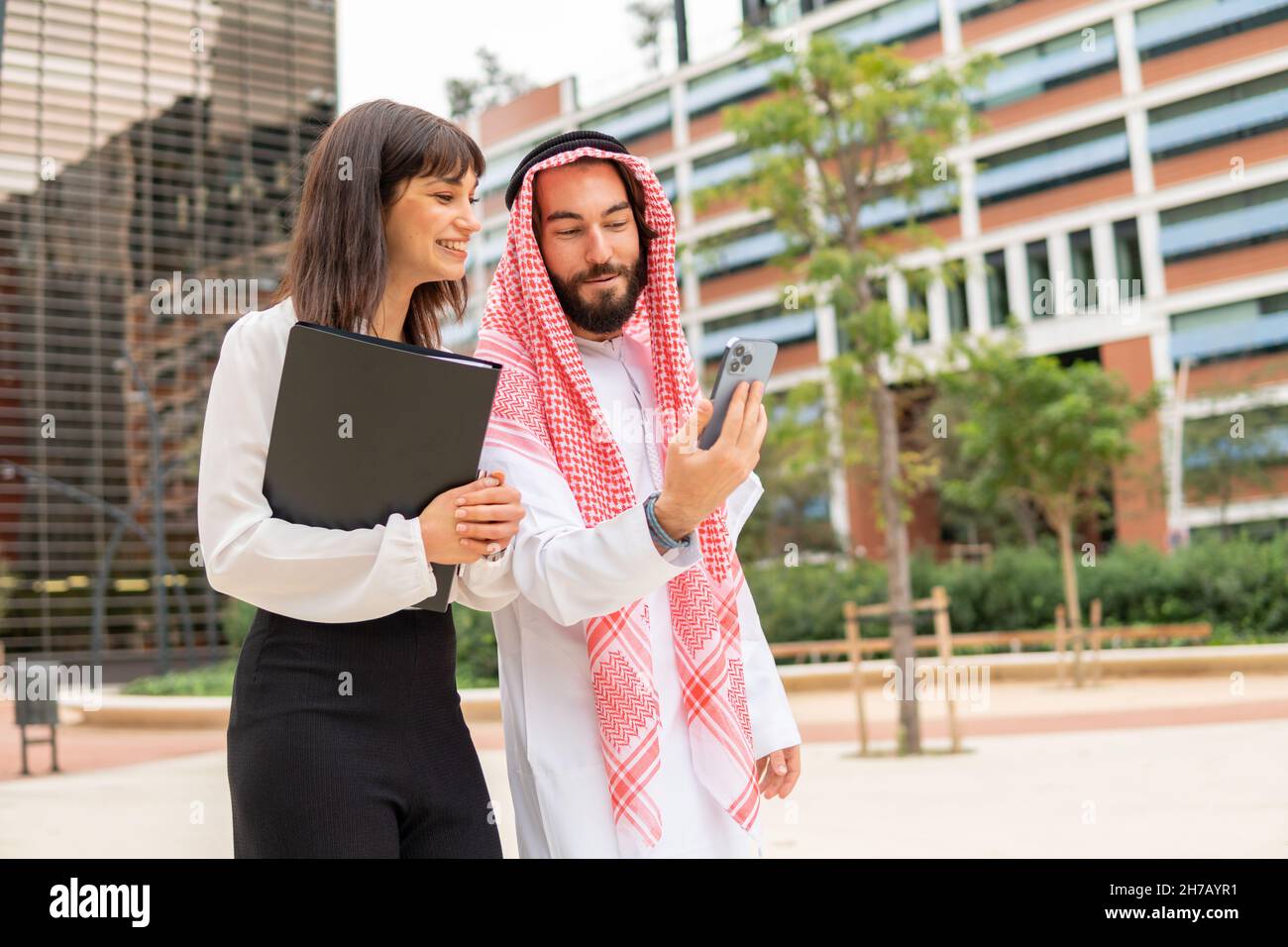 Two diverse business partners arab man and caucasian woman watching video on smartphone during meeting in city, arab man in traditional wear and his female assistant using mobile phone outdoors Stock Photo