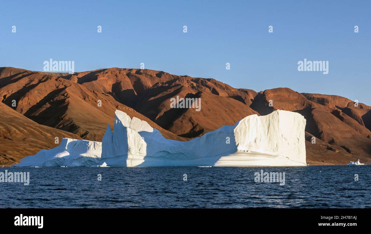 Amazing iron-rich red hills of Rodefjord juxtaposed against a gleaming iceberg, Scoresby Sund, East Greenland Stock Photo