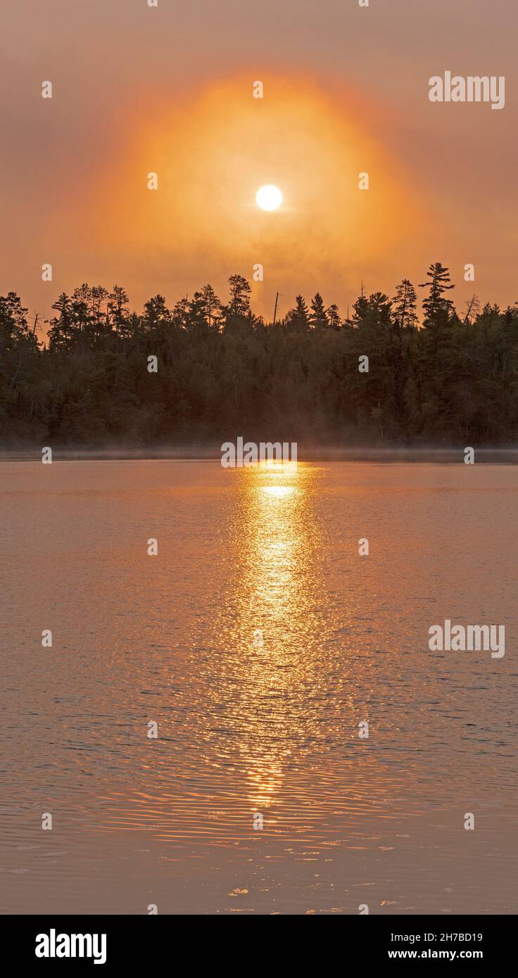 Orange Glow and Reflections of the Rising Sun in Morning Mist on Saganaga Lake in the Boundary Waters in Minnesota Stock Photo