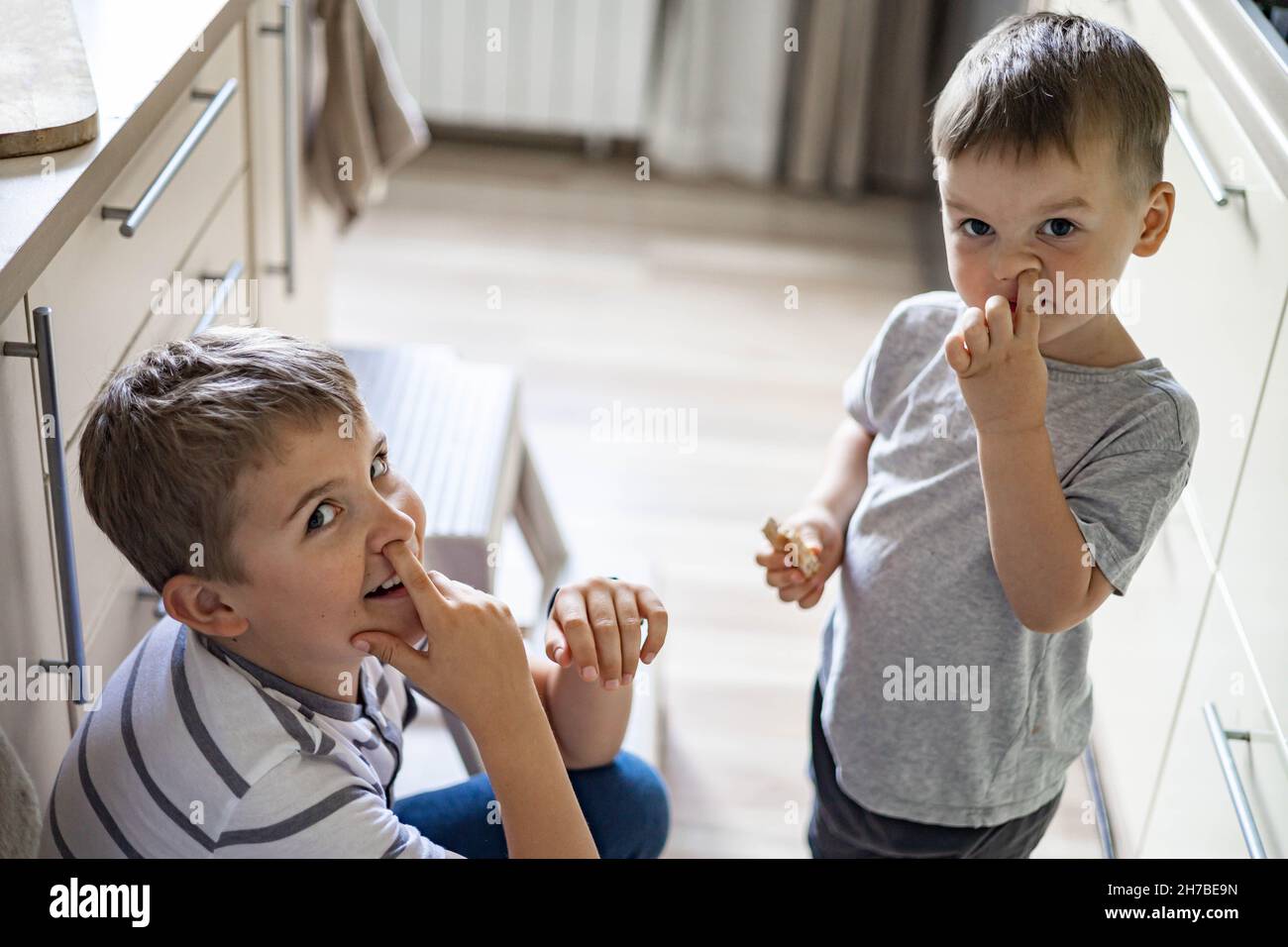 Two funny little boys pick one's nose performing bad habit eating bread at kitchen medium shot Stock Photo