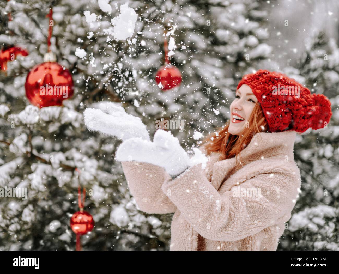 Young smiling beautiful woman in fur coat and mittens standing near decorated Christmas tree and catching snowflakes outdoors Stock Photo