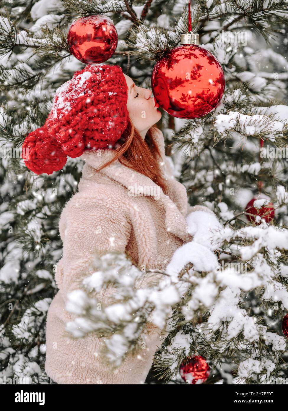 Young woman in fur coat stands outside at decorated Christmas tree and kisses a New Year fir big red ball under snow Stock Photo