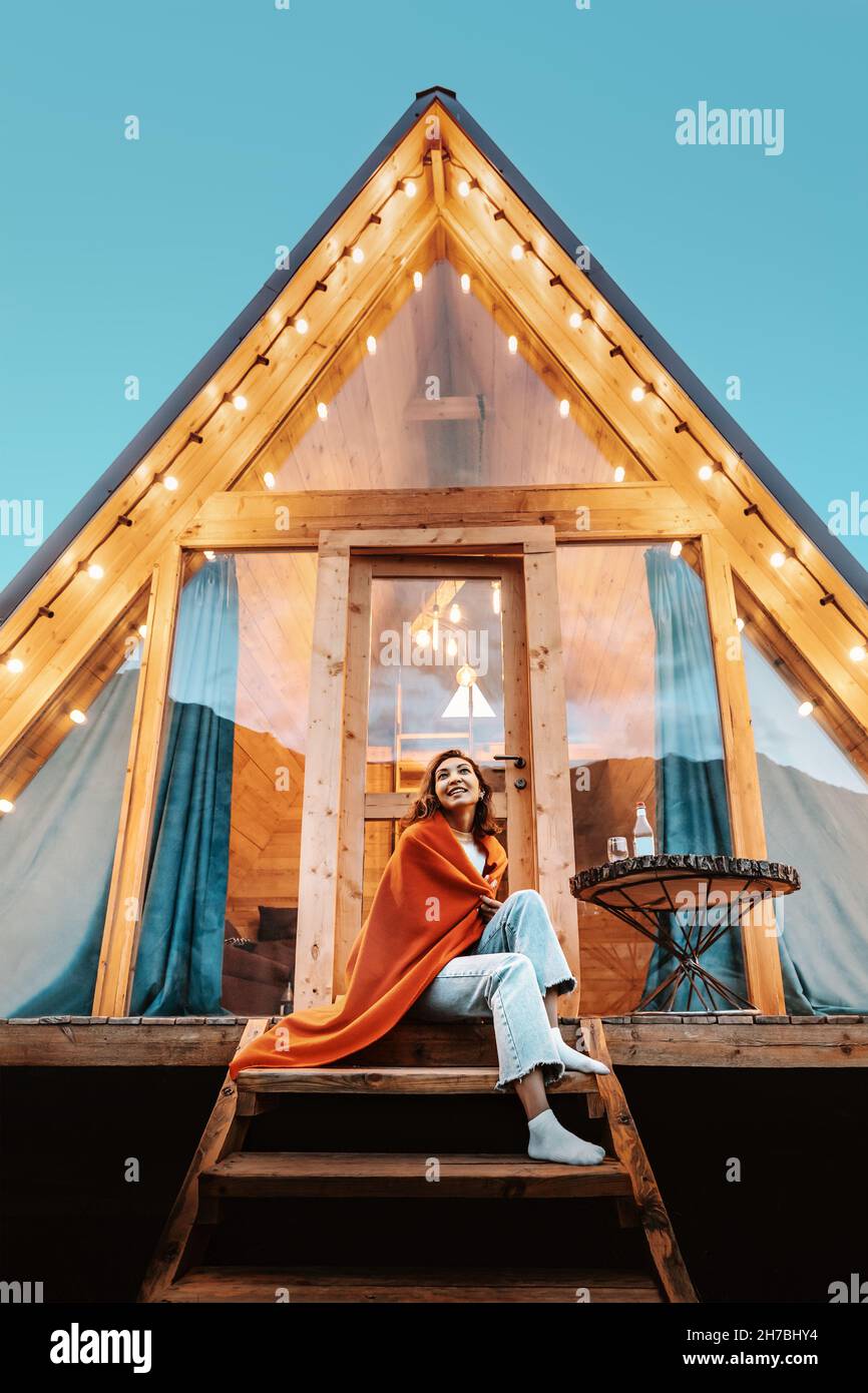 Woman is sitting on the porch of a wooden hut with lights of garlands in the evening. The concept of glamping and renting a chalet. Stock Photo