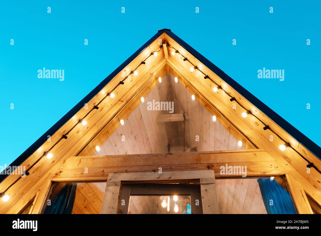 Roof with garlands lights of a small wooden chalet house with glass door and window. The concept of glamping and idyllic holidays Stock Photo