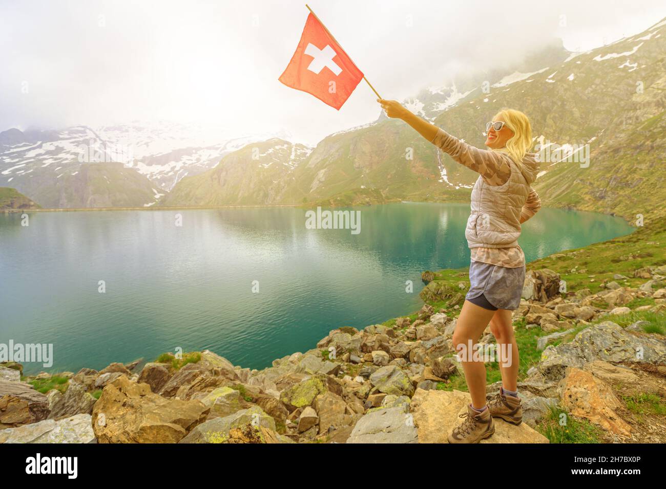 Lady with swiss flag trekking by lakefront of Lake Robiei at sunset. Swiss reservoir in Maggia Valley of Ticino canton. Top of aerial tramway station Stock Photo