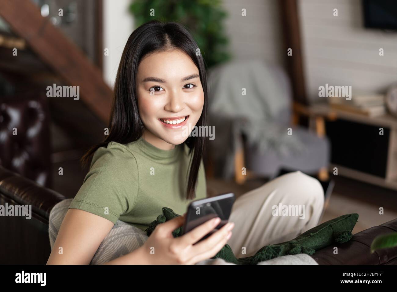 Smiling asian woman sitting on couch at home in cozy clothes, using ...