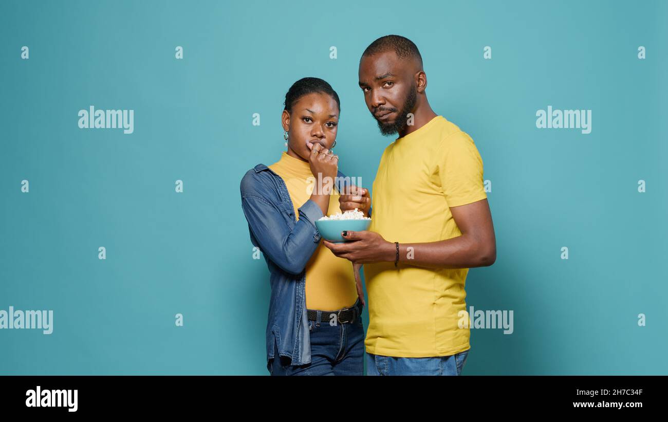 Couple eating popcorn snack from bowl in front of studio camera. Man and woman enjoying movie and watching television, standing over isolated background. People looking at show series. Stock Photo