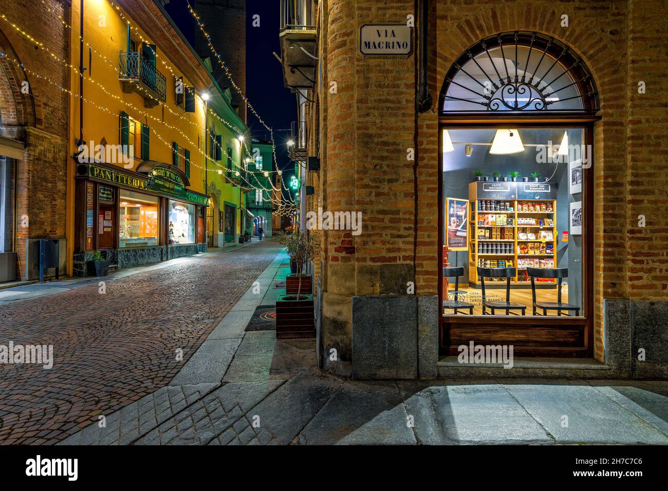 Illuminated window of the coffe shop and narrow cobblestone street with Christmas illumination among historic buildings in Alba, Italy. Stock Photo