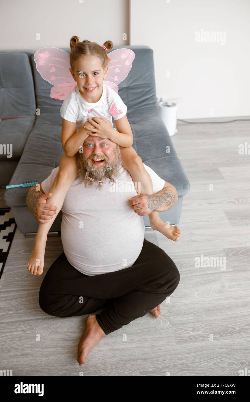 Girl with pink wings sits on shoulders of plump father on floor in living room Stock Photo