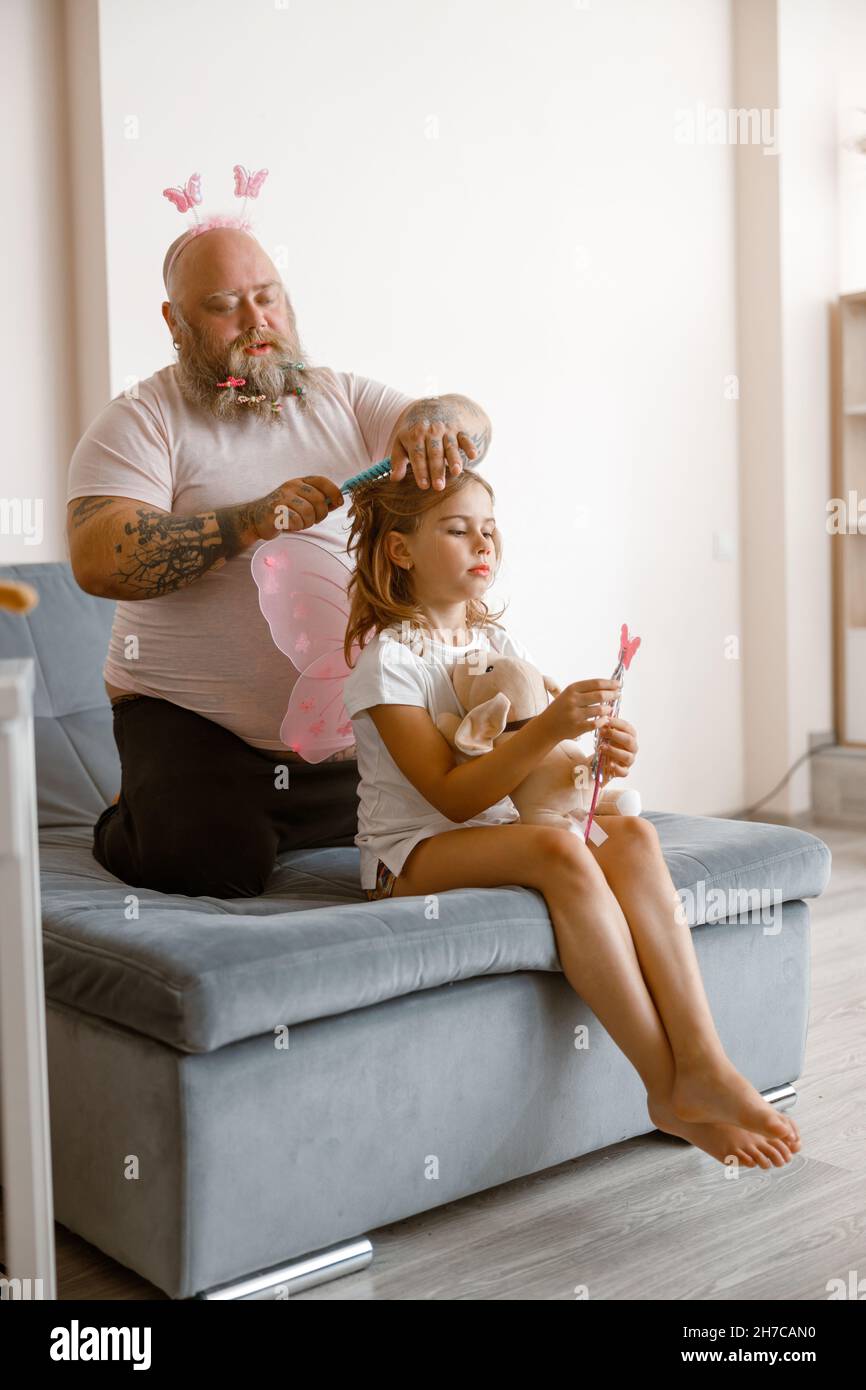 Careful plump father brushes hair of little girl in fairy costume in living room Stock Photo