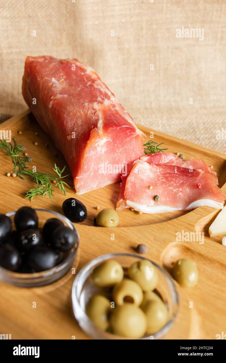 Dry-cured pork fillet on the wooden serving board with cheese, olives, black pepper, tomato sauce. Jerky tenderloin appetizer. Dish of Belarusian Stock Photo