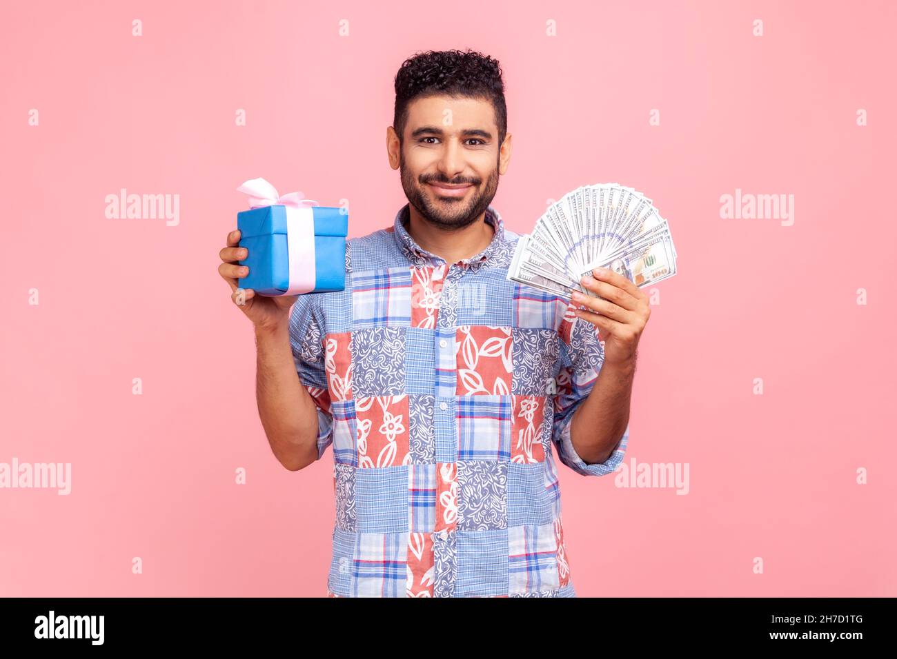 Buying holiday gifts, shopping. Bearded man in blue casual shirt holding wrapped present box and money dollars, looking at camera smiling satisfied. Indoor studio shot isolated on pink background. Stock Photo