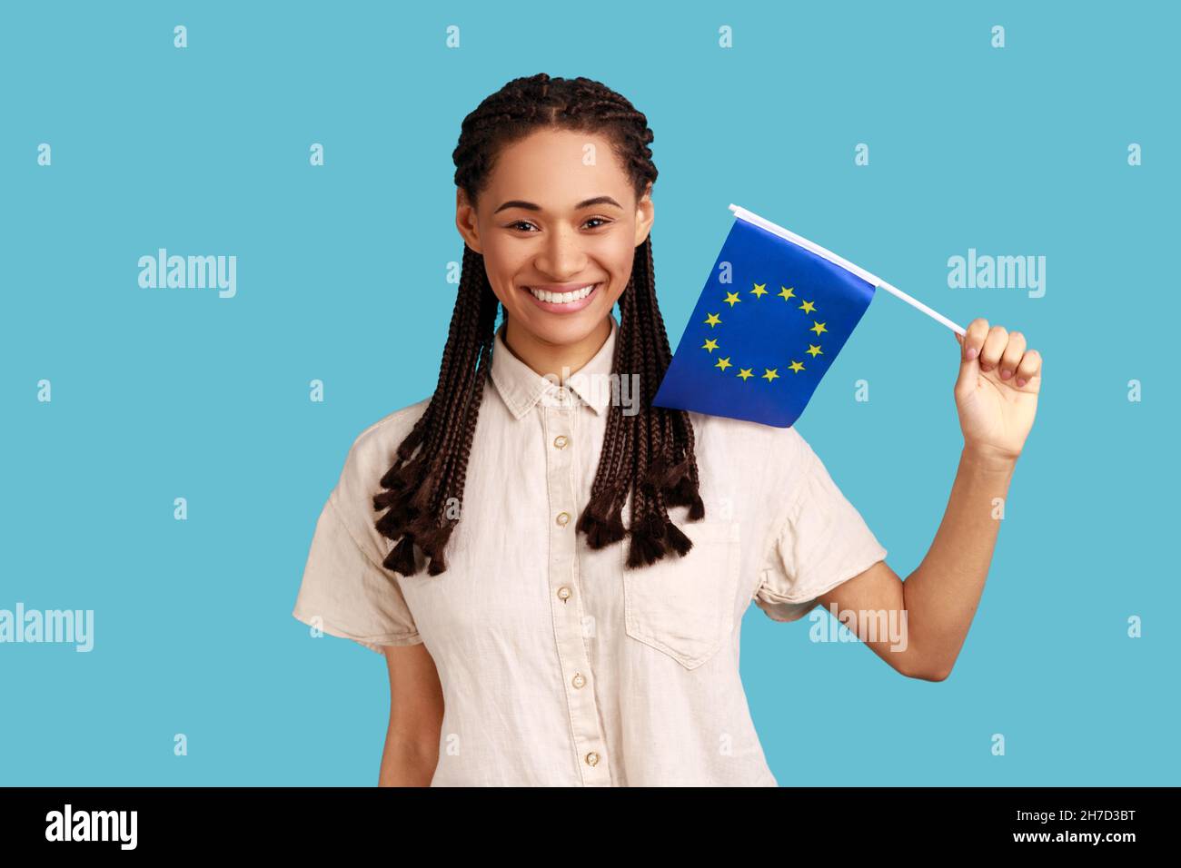 European Union flag. Adorable satisfied smiling woman with black dreadlocks holding Europe flag, looking at camera, wearing white shirt. Indoor studio shot isolated on blue background. Stock Photo